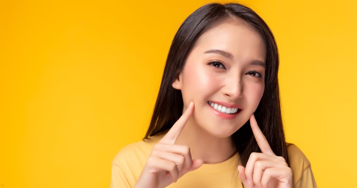 A woman in front of a yellow background, pointing at her mouth and smiling. Her teeth are very white.